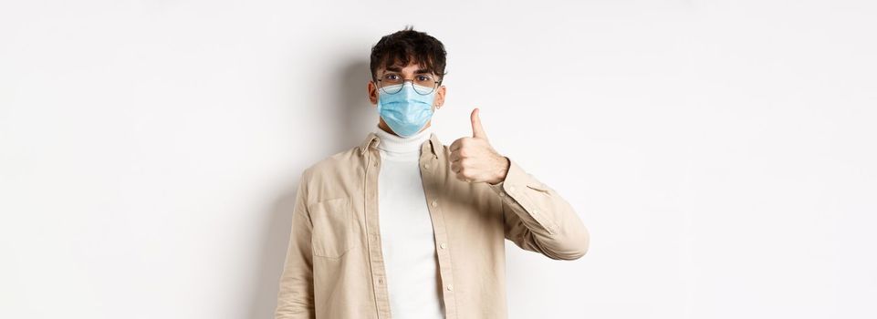 Covid-19, health and real people concept. Satisfied guy in sterile face mask and glasses, showing thumb up in approval, give positive feedback, standing on white background.