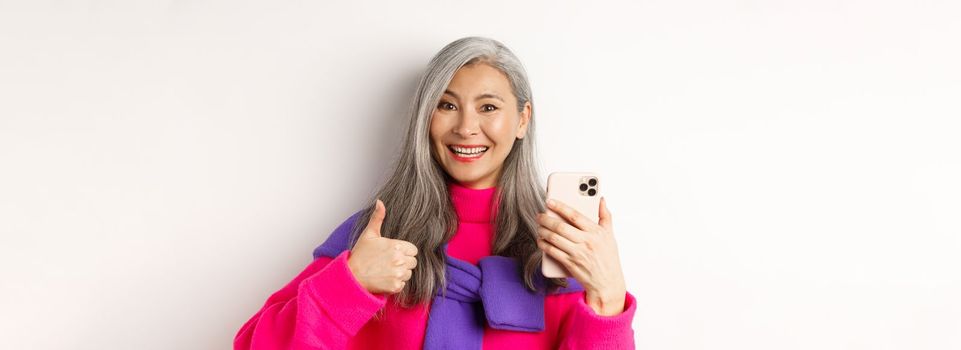 Online shopping. Close up of fashionable asian senior woman showing thumbs-up, using smartphone and approve something, standing over white background.