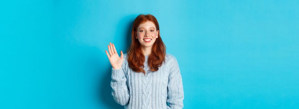 Friendly redhead teenage girl saying hi, waving hand in hello gesture and smiling, standing against blue background.