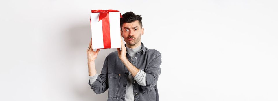 Intrigued young man looking confused at camera, trying to guess what inside gift box from lover, shaking present and stare puzzled, standing on white background. Valentines day concept.