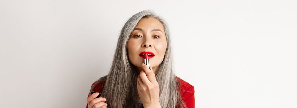 Beauty and fashion concept. Close up of senior asian woman looking in mirror and apply red lipstick, standing over white background.