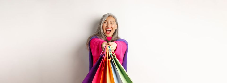 Excited korean lady with grey hair, laughing and showing shopping bags, visit stores with special discounts, standing over white background.