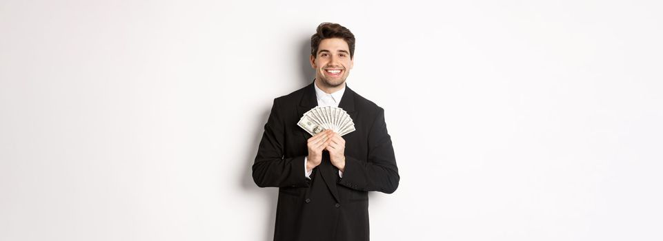 Portrait of happy and pleased handsome man in suit, hugging money and looking satisfied, standing over white background.