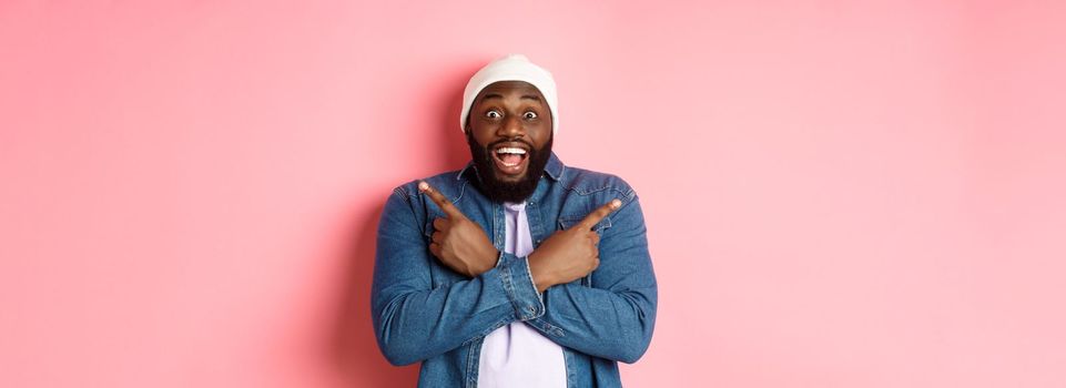 Happy young african-american hipster guy pointing fingers sideways, smiling and showing two choices, showing offers, standing over pink background.