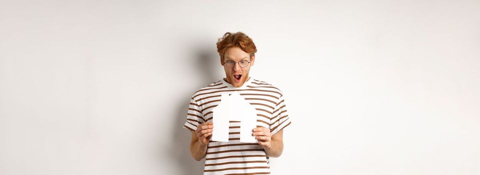 Amazed young man with red hair, staring at paper house cutout with excitement, standing over white background.