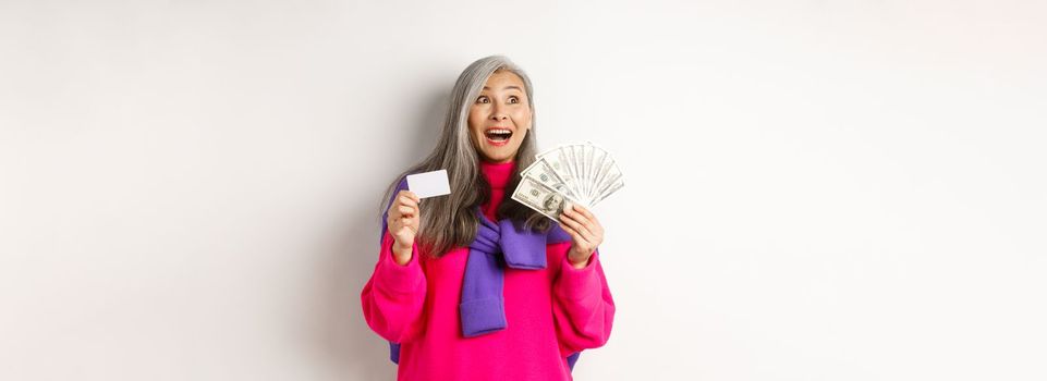 Shopping concept. Happy asian old woman with grey hair, looking fascinated at upper left corner, showing plastic credit card and money dollars, white background.