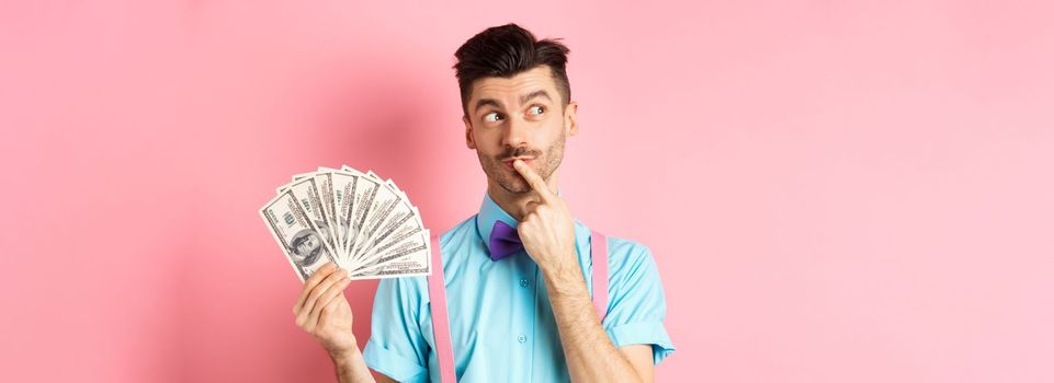 Man thinking about shopping, looking pensive while holding money in dollars, standing over pink background.