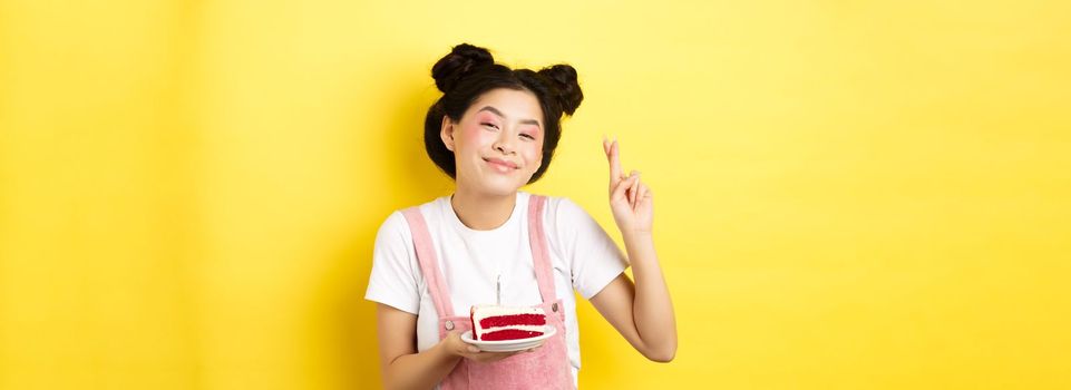 Holidays and celebration. Positive asian birthday girl cross fingers, making wish with b-day cake and lit candle, smiling happy at camera, yellow background.