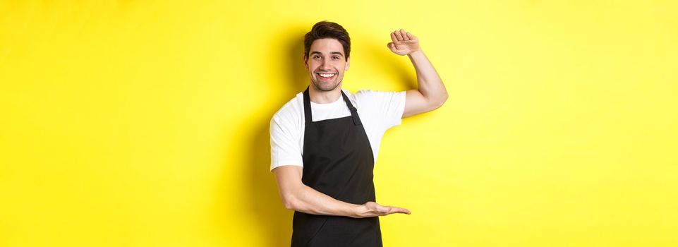 Smiling handsome barista showing something long or large, standing over yellow background.