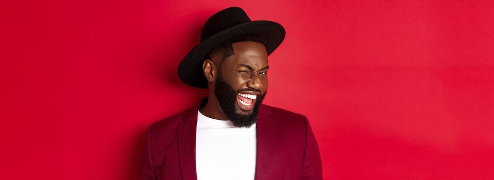 Cheeky african american man in party outfit, winking at camera and smiling, standing against red background.