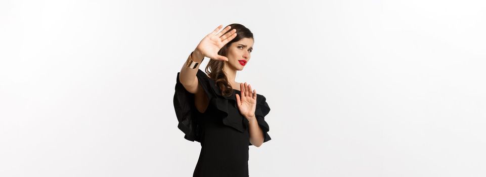 Fashion and beauty. Reluctant and worried woman asking to stay away, showing stop gesture and looking scared, standing in black dress over white background.