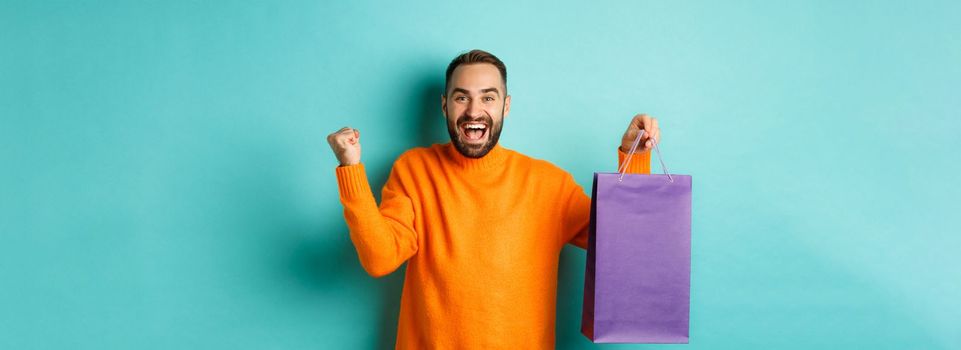 Happy man holding purple shopping bag and rejoicing, got discount and celebrating, standing over turquoise background.