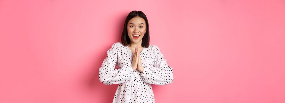 Beautiful asian woman thanking you, holding hands together in appreciation gesture, smiling happy at camera, standing grateful over pink background.