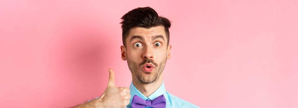 Close-up of funny guy with moustache, say wow and showing thumb up in approval, checking out something cool, recommending product, standing on pink background.