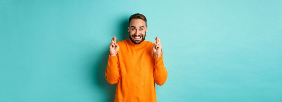 Excited man with beard, making a wish, holding fingers crossed for good luck and smiling, standing over light blue background.
