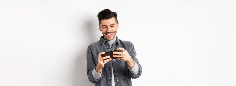 Handsome young man playing mobile video game and smiling at screen, standing against white background.