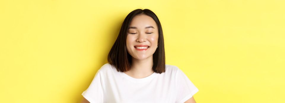 Close up of happy, romantic asian girl dreaming of something, close eyes and smiling delighted, standing over yellow background.