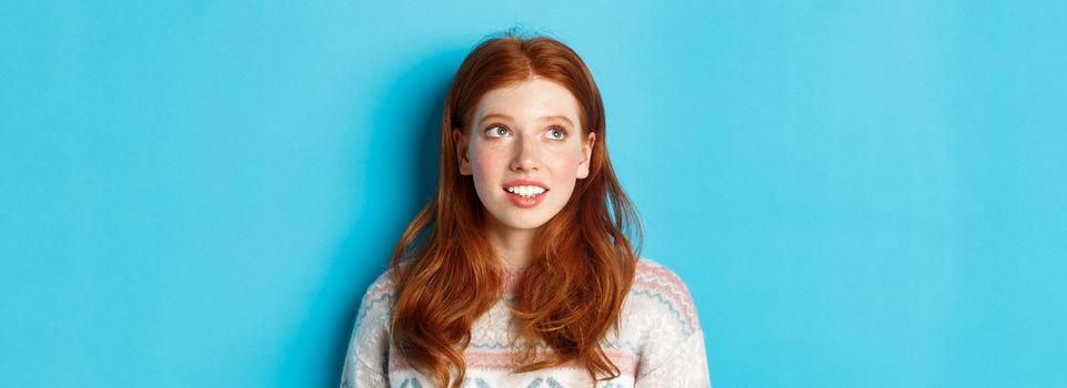 Close-up of dreamy teen girl with red hair, looking at upper left corner and smiling, standing against blue background.