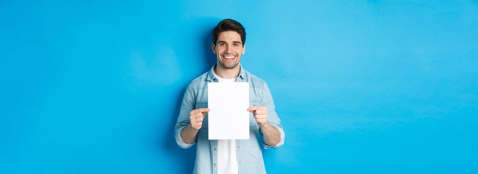 Young smiling guy in casual outfit, holding blank piece of paper with your advertisement, standing over blue background.