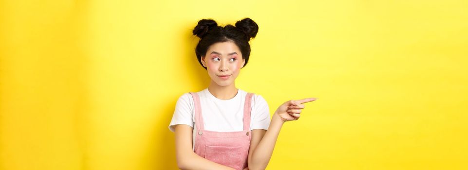 Cute beauty girl with makeup and summer clothes, pointing and looking left at promo banner, standing curious on yellow background.