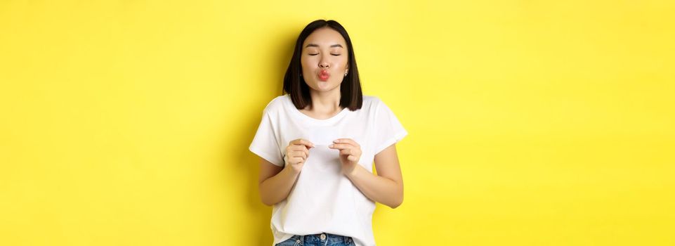 Young asian woman in casual white t-shirt showing plastic credit card and pucker lips silly, going on shopping, yellow background.