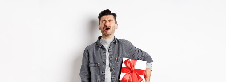 Valentines day heartbreak. Single and heartbroken guy crying lonely, holding big gift box, being rejected by lover, sobbing and feeling alone, standing on white background.