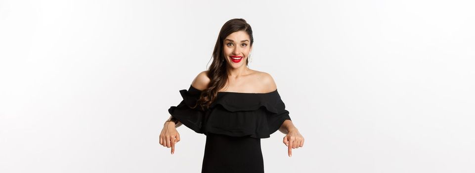 Fashion and beauty. Elegant woman in black dress pointing fingers down, showing promo and smiling, standing over white background.