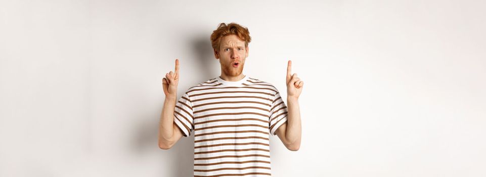 Confused young man with red curly hair pointing fingers up, staring shocked and puzzled, standing over white background.