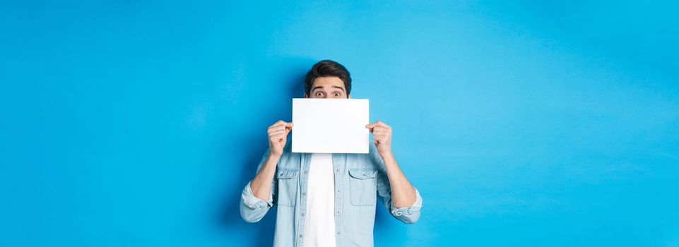 Excited man hiding face behind blank paper, place for your logo, standing over blue background.