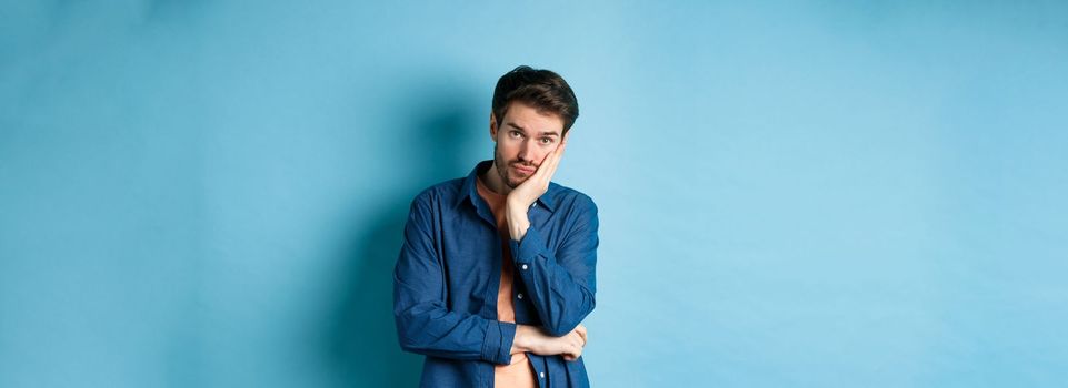 Sad and bored man lean face on hand, looking at camera empty-headed, standing bothered on blue background.