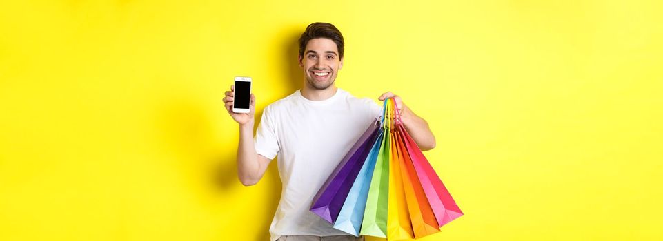 Young man holding shopping bags and showing mobile phone screen, money application, standing over yellow background.