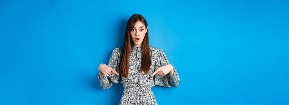 Excited young woman in dress pointing fingers down at logo, looking intrigued at camera, standing on blue background.