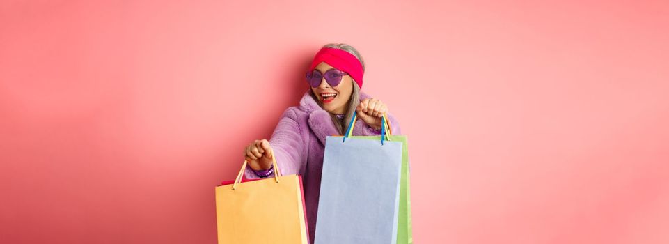 Funky and cool asian senior woman in fashionable clothes dancing while going shopping on sales, holding shop paper bags and having fun, pink background.