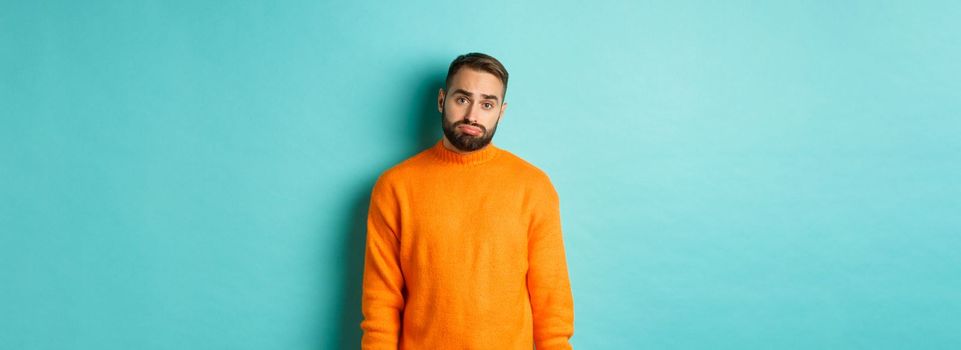 Sad man looking at camera, sulking and feeling distressed, standing over light blue background.