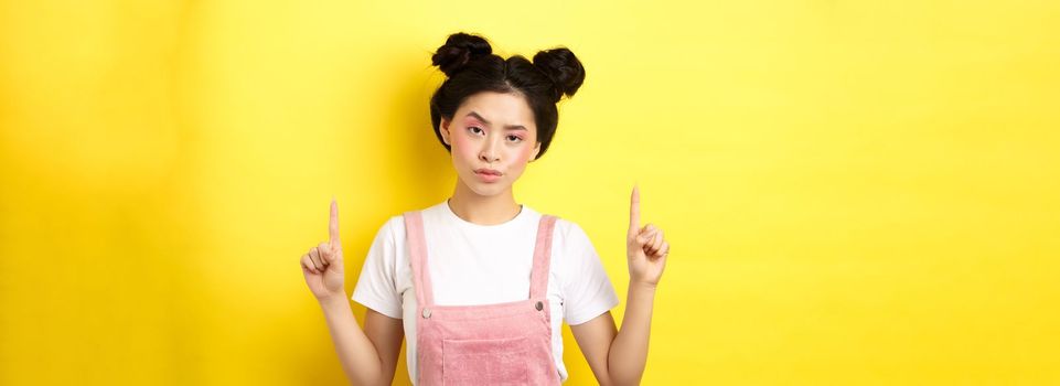 Annoyed and tired asian girl with pink glamour makeup, pointing fingers up and look bothered, standing on yellow background.