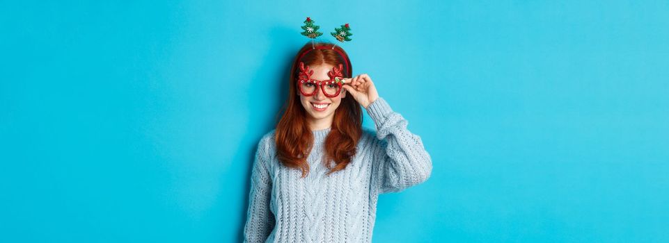 Winter holidays and Christmas sales concept. Beautiful redhead female model celebrating New Year, wearing funny party headband and glasses, smiling at camera.