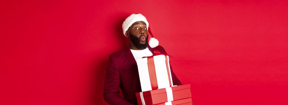 Christmas, New Year and shopping concept. Happy Black man in santa hat and blazer holding xmas presents, bring gifts and smiling, standing against red background.