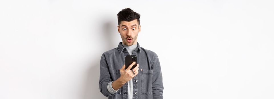 Man look surprised at smartphone screen, receive exciting news on phone, standing against white background.