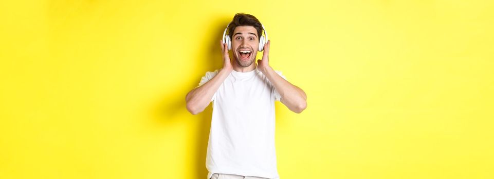 Man in headphones looking surprised and happy, listening awesome song, standing over yellow background.