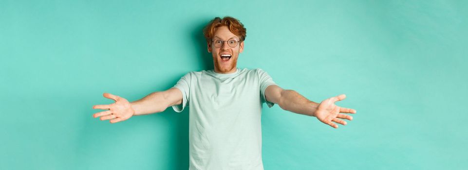 Excited young redhead guy in glasses strethced out hands in warm welcome, invite you and smile friendly at camera, standing happy over turquoise background.