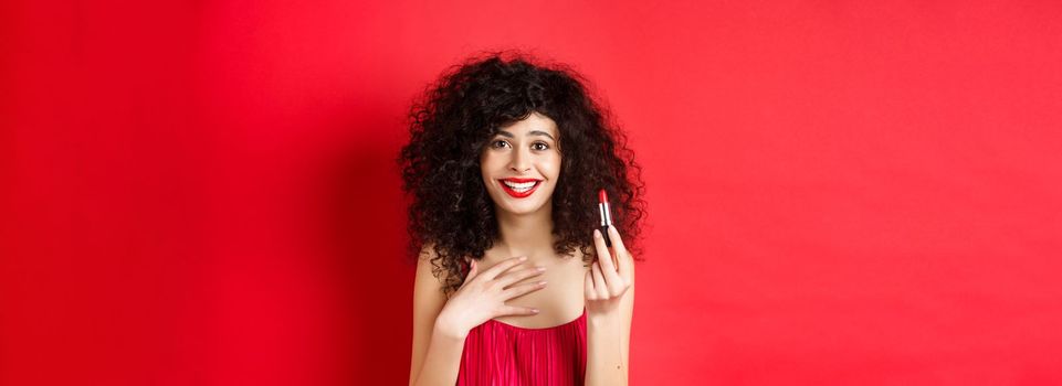 Beauty and make up concept. Beautiful female model with curly hair, wearing evening dress, showing red lipstick and smiling, standing on white background.