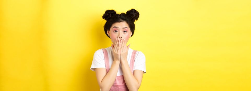 Shocked asian girl gasping amazed, staring at something shocking, covering mouth with hands, standing with beauty makeup and summer clothes on yellow background.