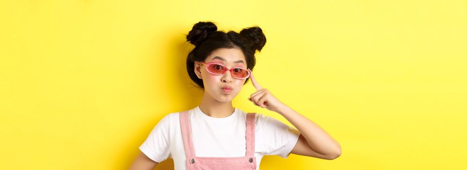 Stylish teen asian girl wearing sunglasses and pink overalls, standing on yellow background.