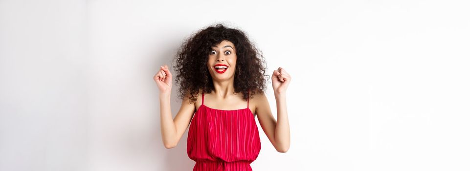 Excited curly-haired woman with evening makeup, gasping fascinated, raising hands up and rejoicing, winning prize and celebrating, standing against white background.