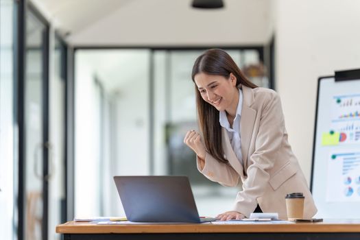 Successful young Asian business woman achieving goals excited raised hands rejoicing with laptop in the office. New startup project concept.