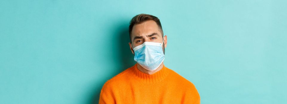 Covid-19, social distancing and quarantine concept. Upset man in face mask complaining, looking displeased and tired, standing against light blue background.