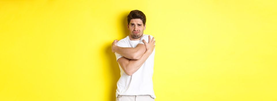 Silly and timid guy trying to comfort himself, hugging his body and frowning scared, standing over yellow background.