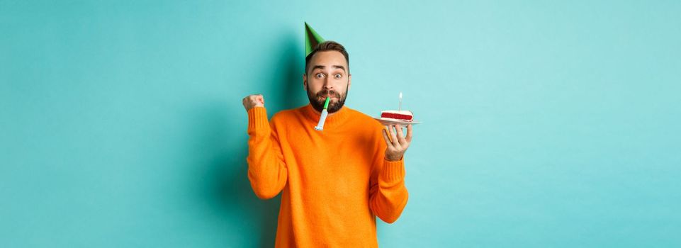 Happy birthday guy celebrating, wearing party hat, blowing wistle and holding bday cake and doing fist pump excited, standing over turquoise background.