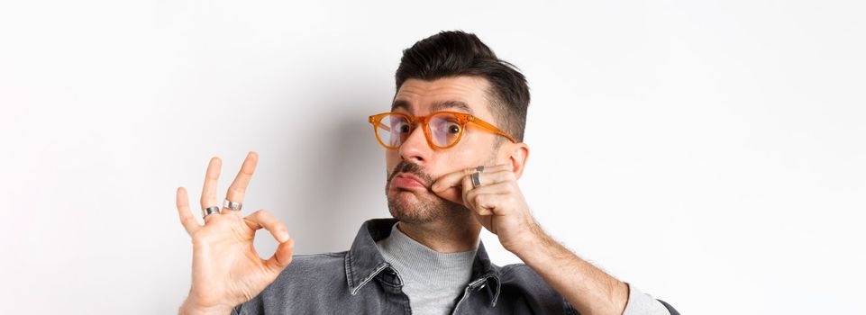 Funny young man in glasses touching his french moustache and showing okay, not bad sign, approve and like good product, standing on white background.