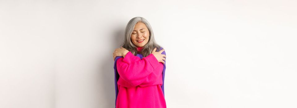 Valentines day and holidays concept. Lovely asian senior woman in pink sweater hugging herself with eyes closed, smiling, standing over white background.